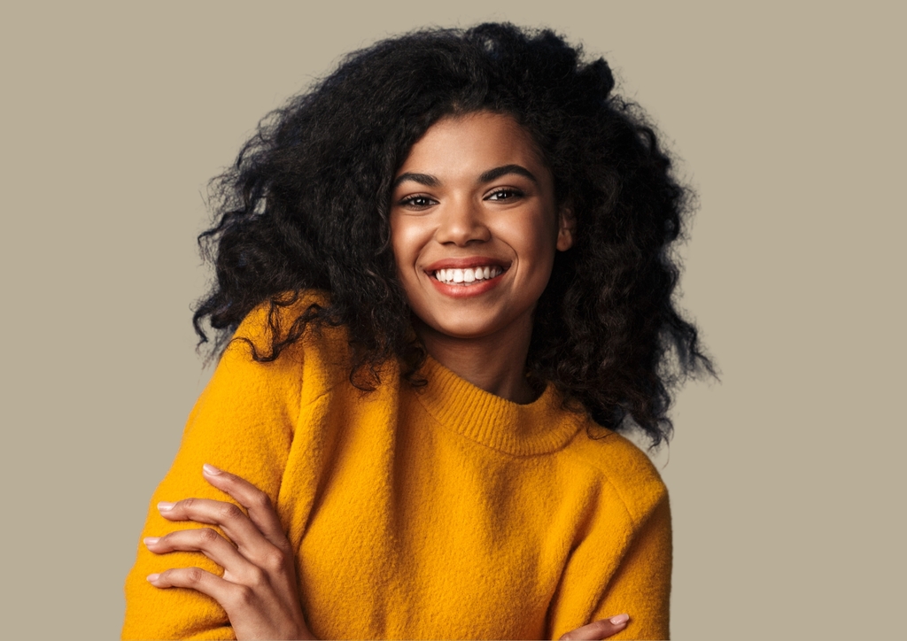 A person with long, curly black hair smiles warmly at the camera. They are wearing a bright yellow sweater and stand against a plain, neutral background, exuding an aura of cultural and linguistic diversity. Their arms are crossed loosely, contributing to a relaxed and cheerful demeanor.