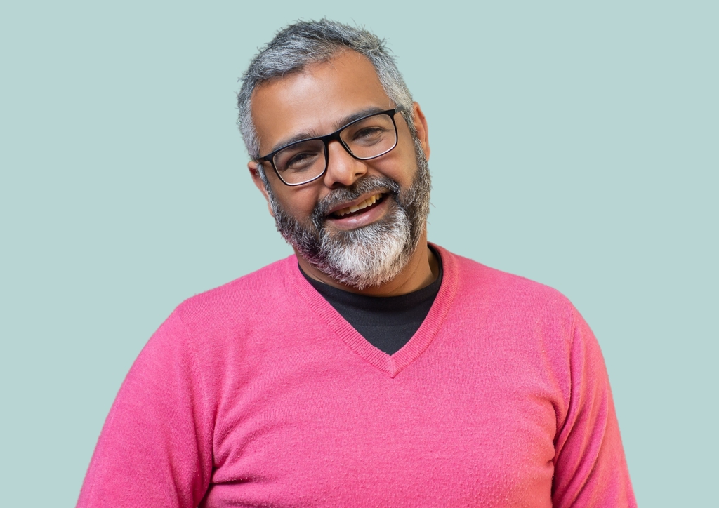 A middle-aged man with short gray hair and a full beard smiles warmly at the camera in Hervey Bay. He is wearing black-framed glasses, a bright pink V-neck sweater over a black shirt, set against a plain light gray background, embodying the vibrant spirit of this town known for its affordable housing.