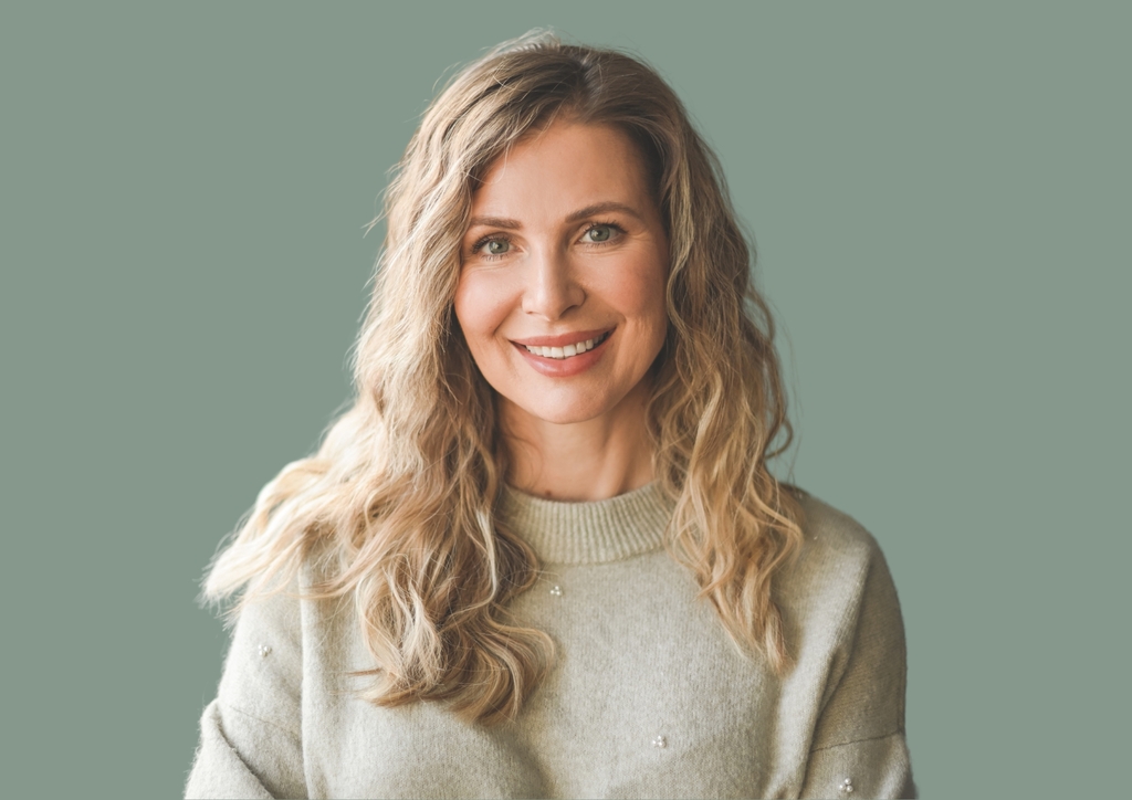 A woman with long, wavy blond hair smiles warmly, framed by a muted green background that echoes the serene ambiance of Shorelyne. She wears a light gray sweater with small embellishments, reflecting the calm and cozy essence of secure rentals.